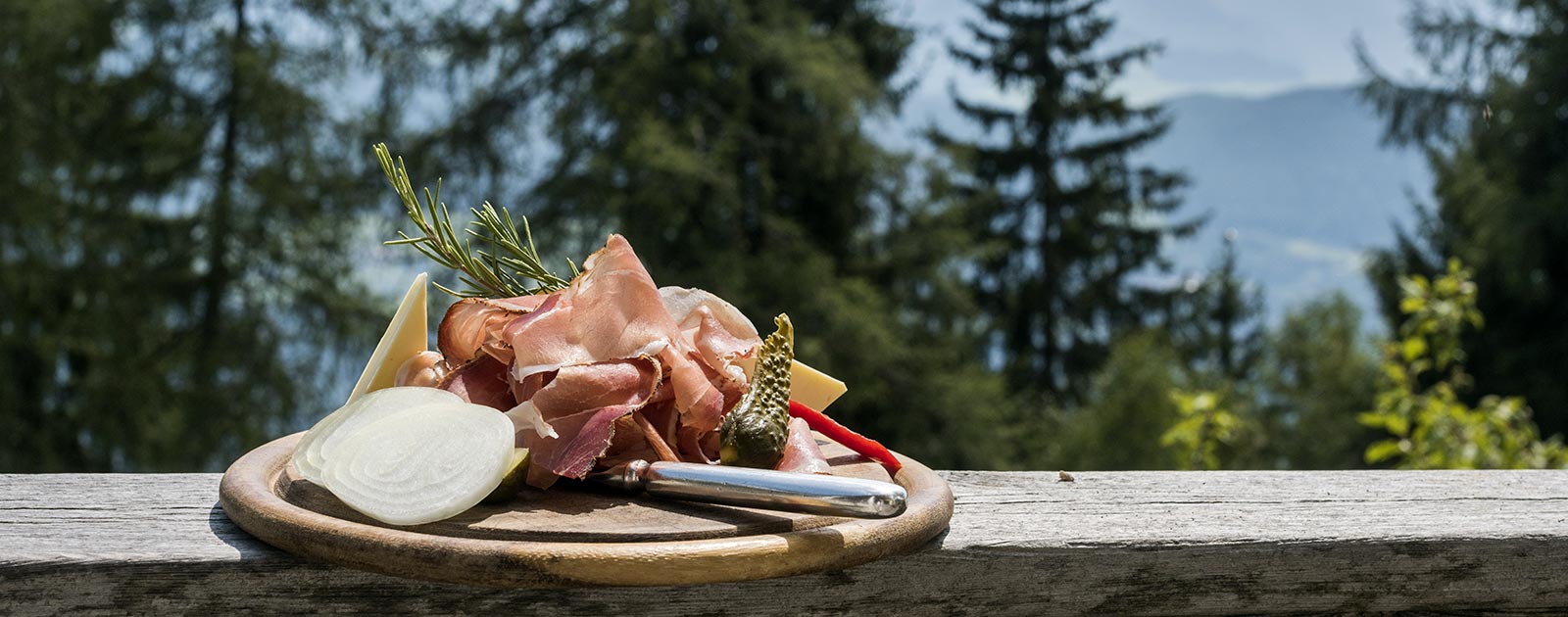 ein traditionelles Speckbrettl mit sauren Gurken, Käse, Zwiebeln und Rosmarin auf der Terrasse des Briol im Sommer